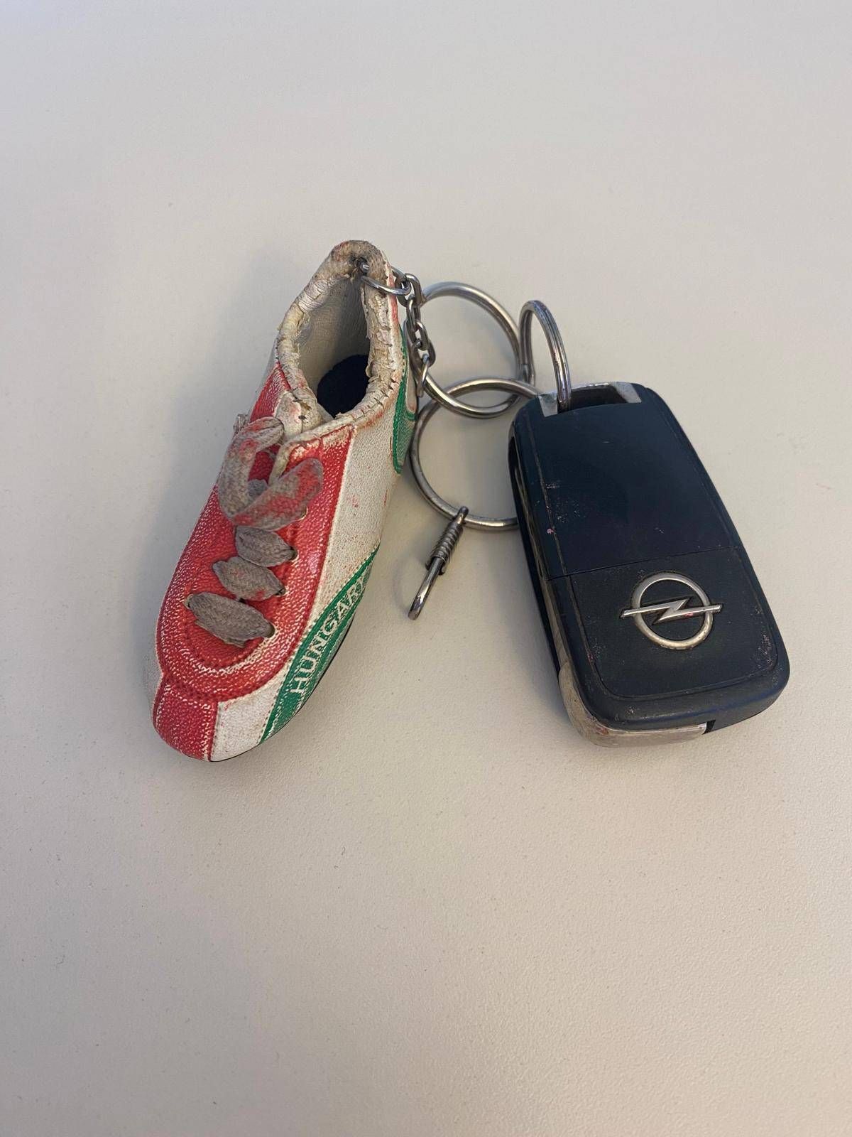 A small shoe-shaped keychain with a red, white, and green design, attached to a silver keyring, and a black car key with a silver Opel logo.