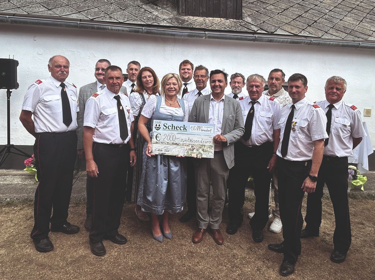 A group of people dressed in uniforms are standing in front of a building, with a woman holding a large check and a man standing next to her.