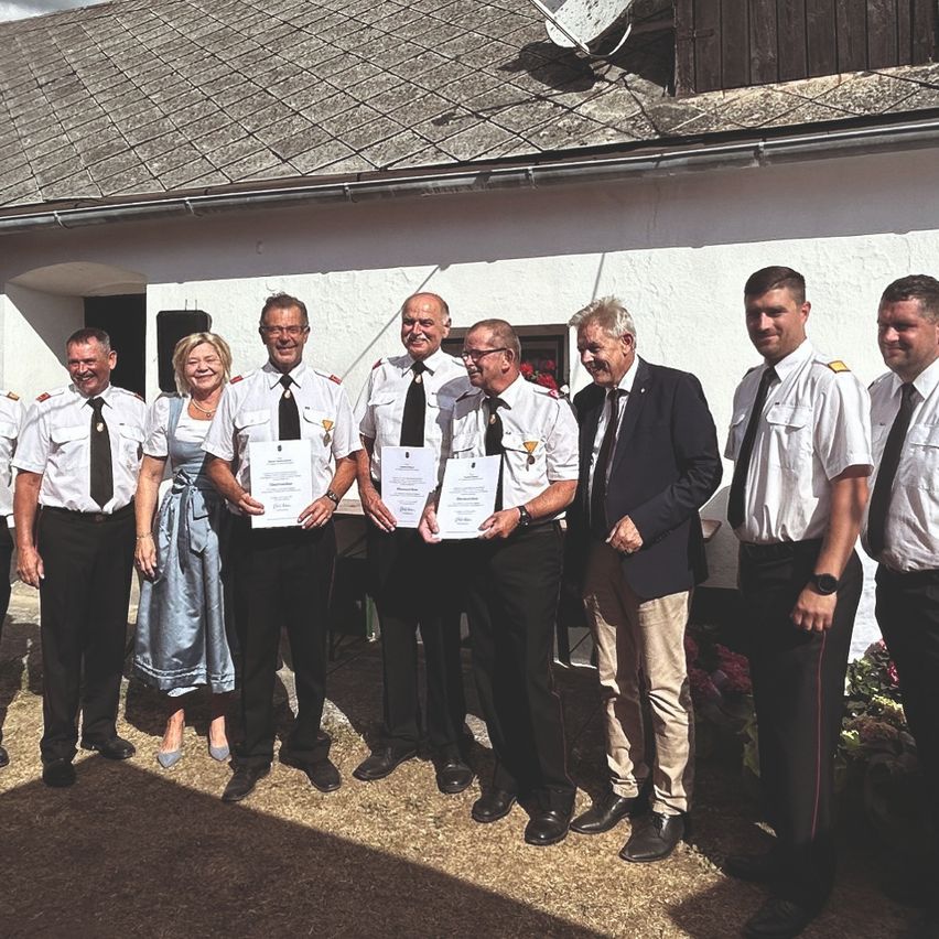 A group of people dressed in uniforms stand outside a building, holding certificates. The building has a satellite dish on its roof.