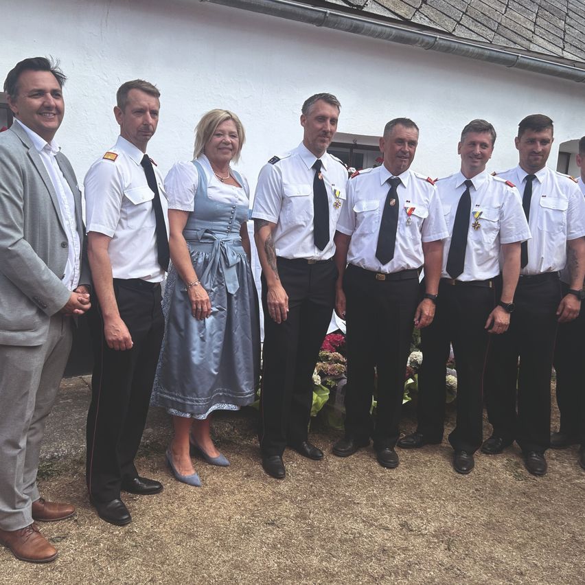 A group of people dressed in uniforms and formal attire stand in front of a white building with flowers and greenery in the background.