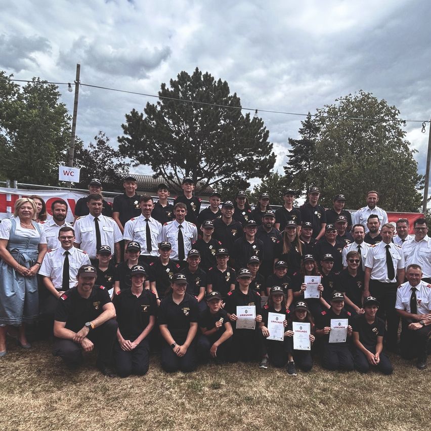 A group of people in uniform, some holding certificates, pose for a photo in an outdoor setting with trees and a cloudy sky in the background.