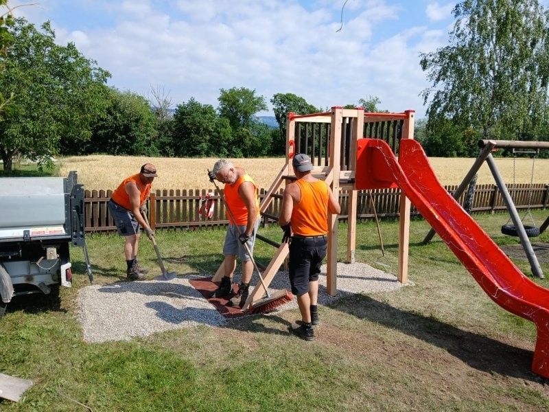 Three men in orange vests are cleaning a playground area with a red slide and a wooden structure. A truck is parked nearby.