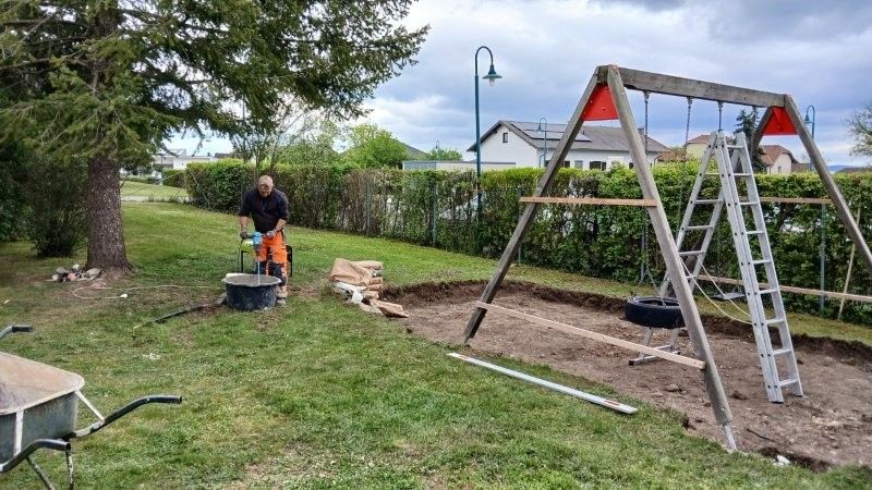 A man is working on a backyard project, standing near a wheelbarrow and a swing set with a tire swing. He is wearing gloves and holding a tool.