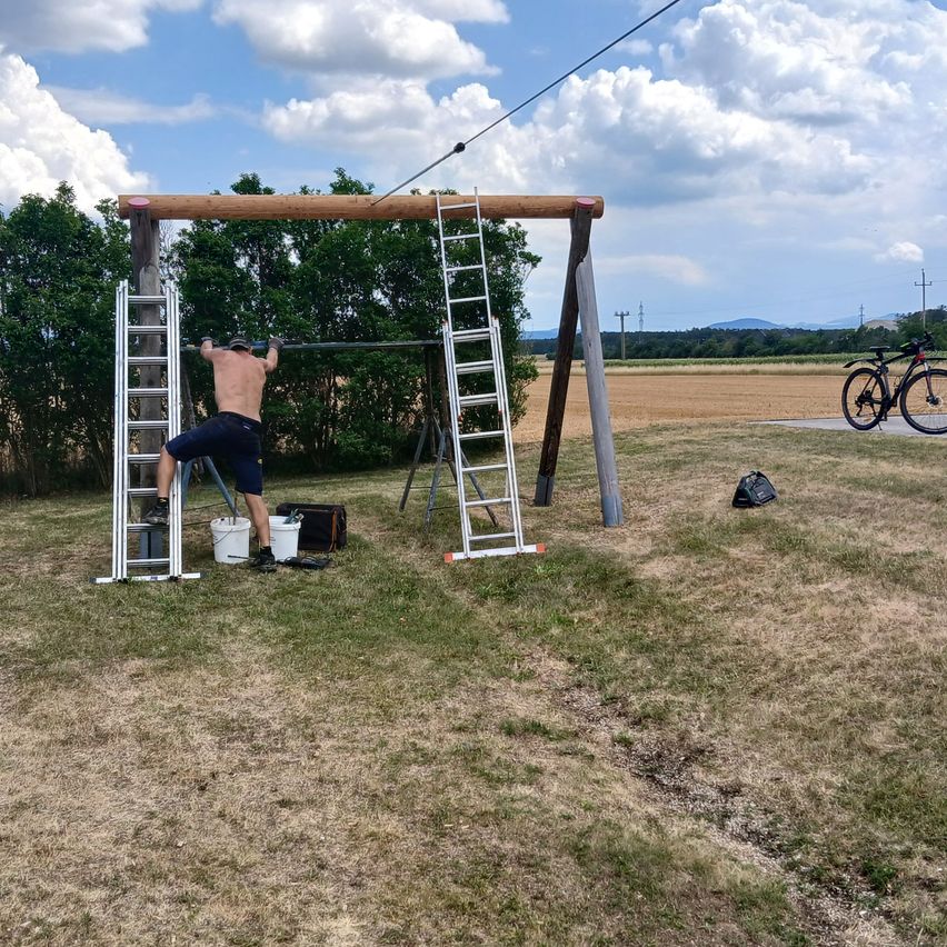 A man is climbing a wooden structure with a ladder on each side, holding onto the top bar. He is shirtless and wearing shorts.