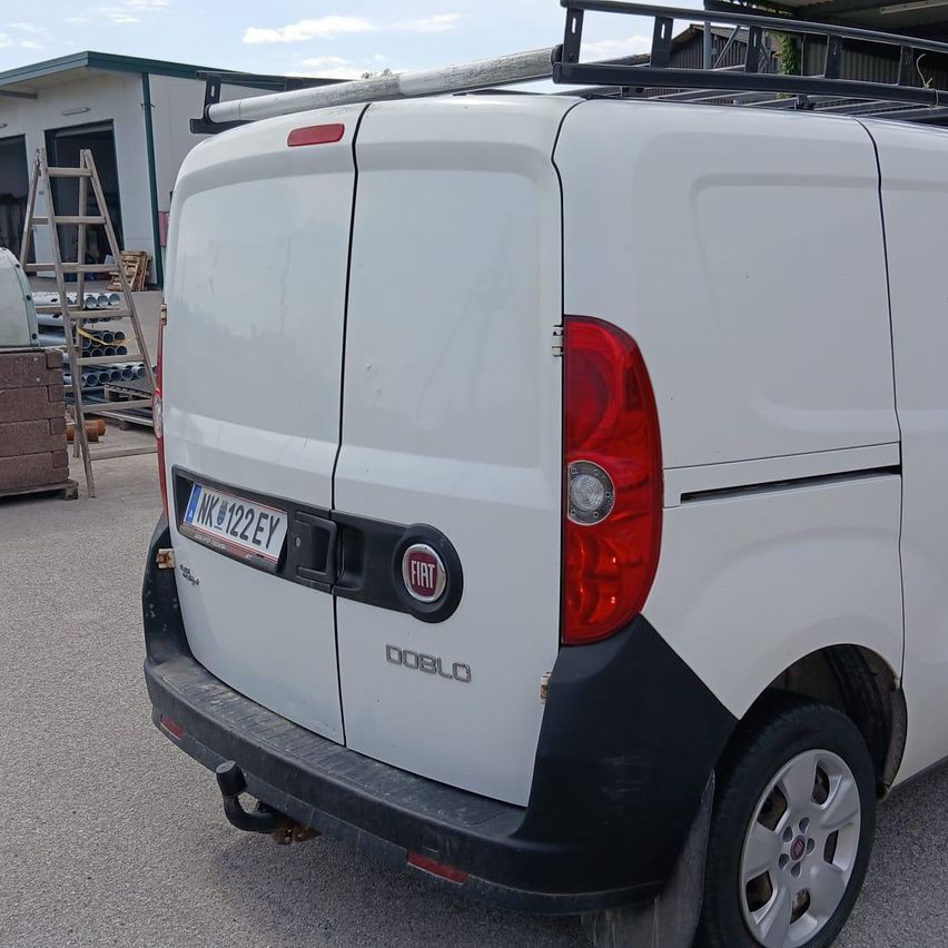 Die Heckansicht eines weißen Fiat Doblo Lieferwagens mit Dachträger, geparkt in einem Außenbereich mit einem Gebäude und einer Leiter im Hintergrund.