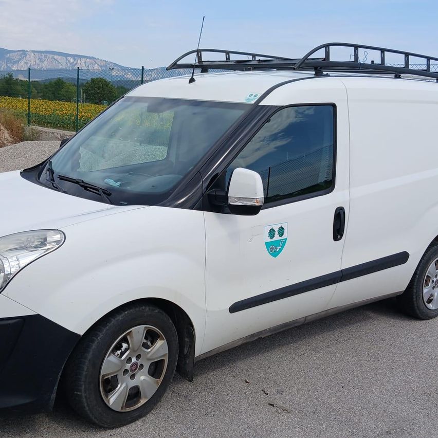 Ein weißer Lieferwagen mit Dachträger ist auf einer gepflasterten Oberfläche geparkt. Er hat ein Logo an der Seitentür und einen grünen Feld- und Berghintergrund.