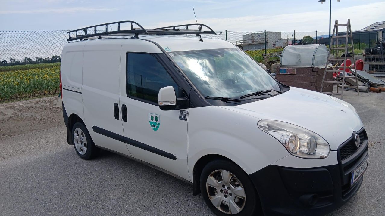 Ein weißer Lieferwagen mit einem grünen Logo auf der Tür, geparkt auf einer Betonfläche. Er hat ein Dachgepäckträger und eine silberne Felge am Vorderreifen.