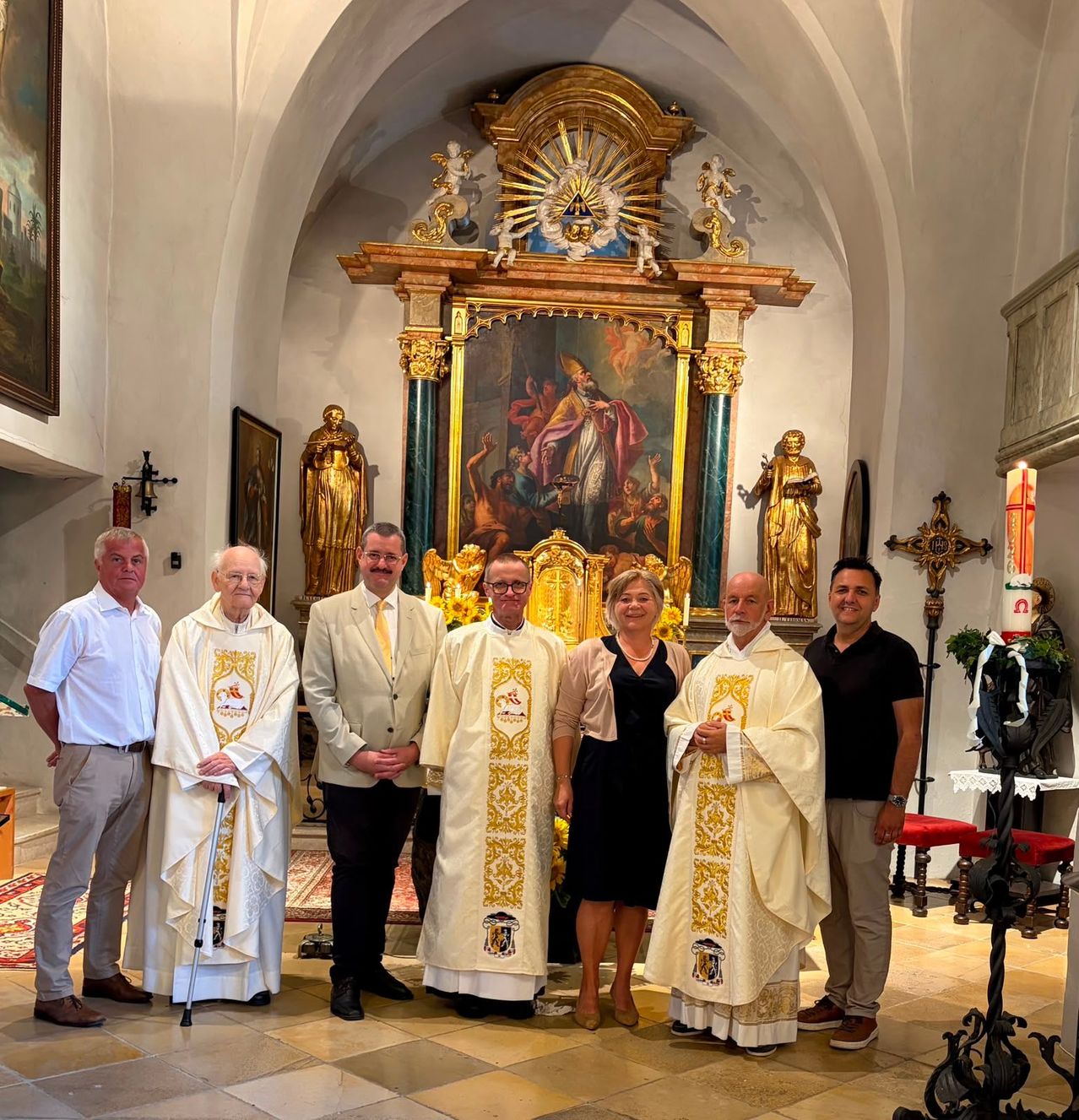 Eine Gruppe von Personen, darunter zwei Priester, steht vor einem kunstvollen Altar in einer Kirche und posiert für ein Foto.