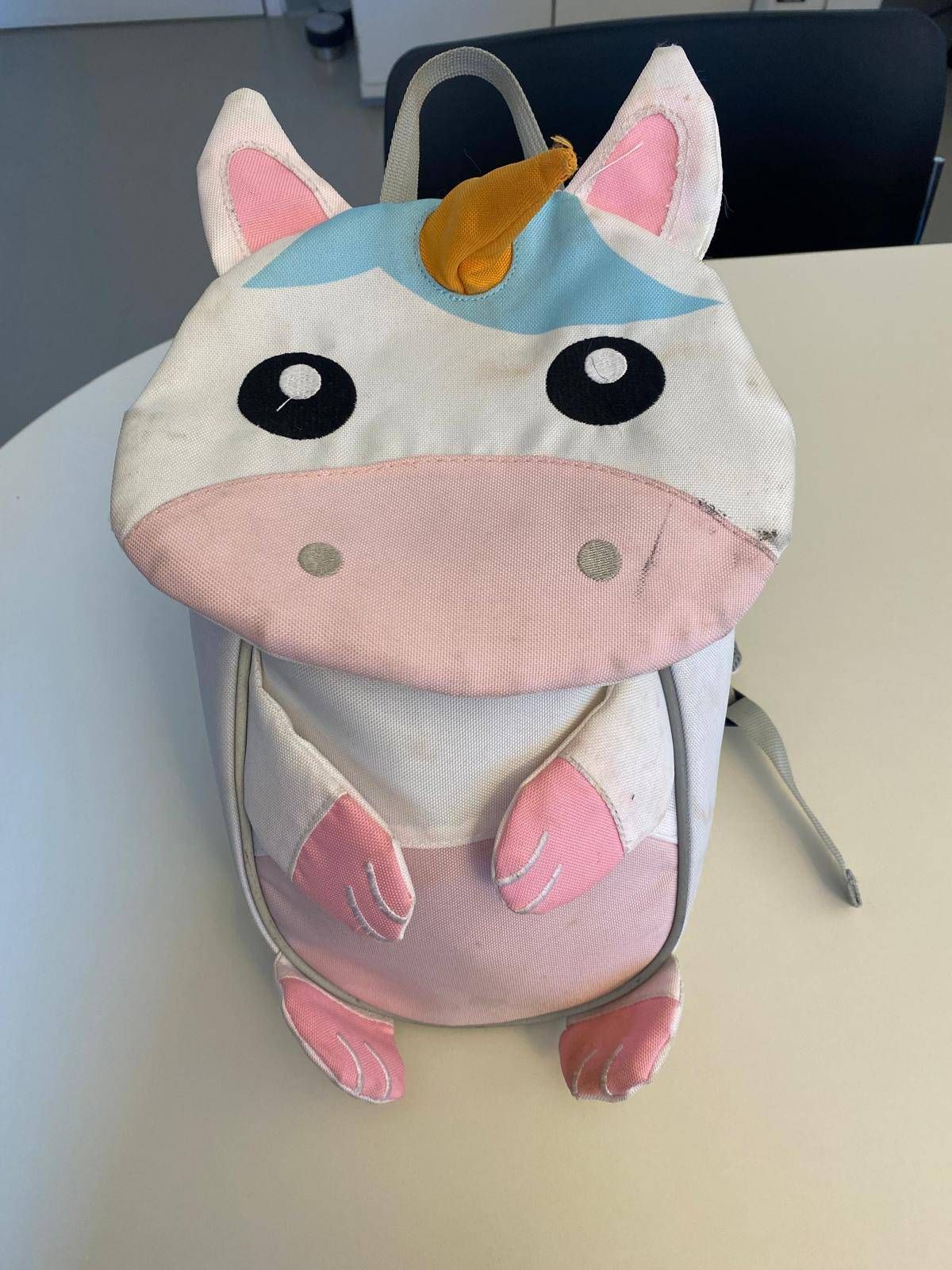 A pink and white backpack shaped like a unicorn with a blue horn and large black eyes. The backpack is placed on a white surface.