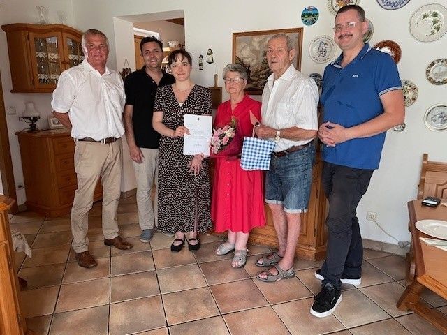 A group of people standing in a room, a woman holding a certificate and flowers, and a man holding a bag.