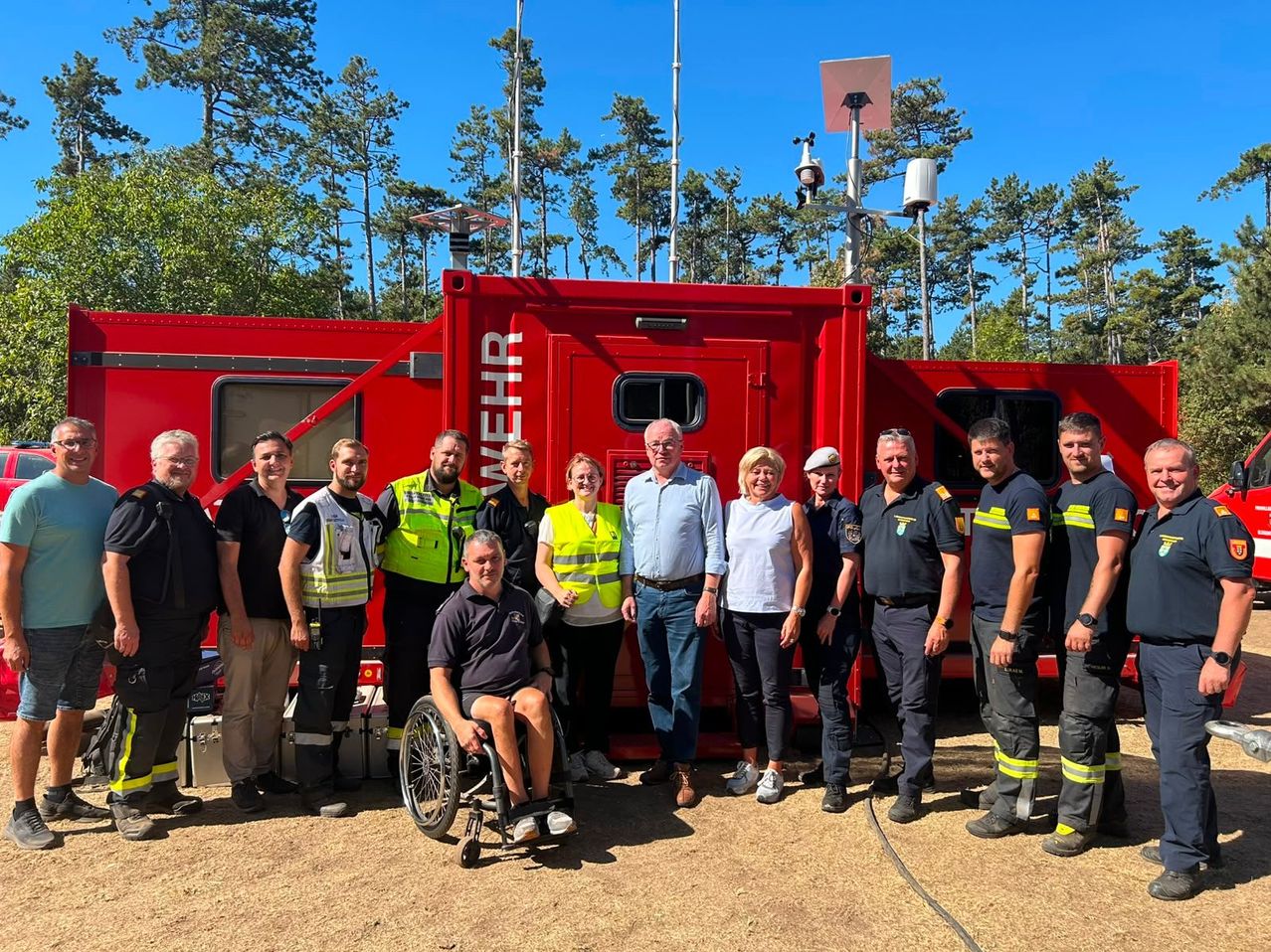A group of people in uniforms and safety vests stand in front of a red vehicle with the word 'wehr' on it. Trees and a clear blue sky are in the background.
