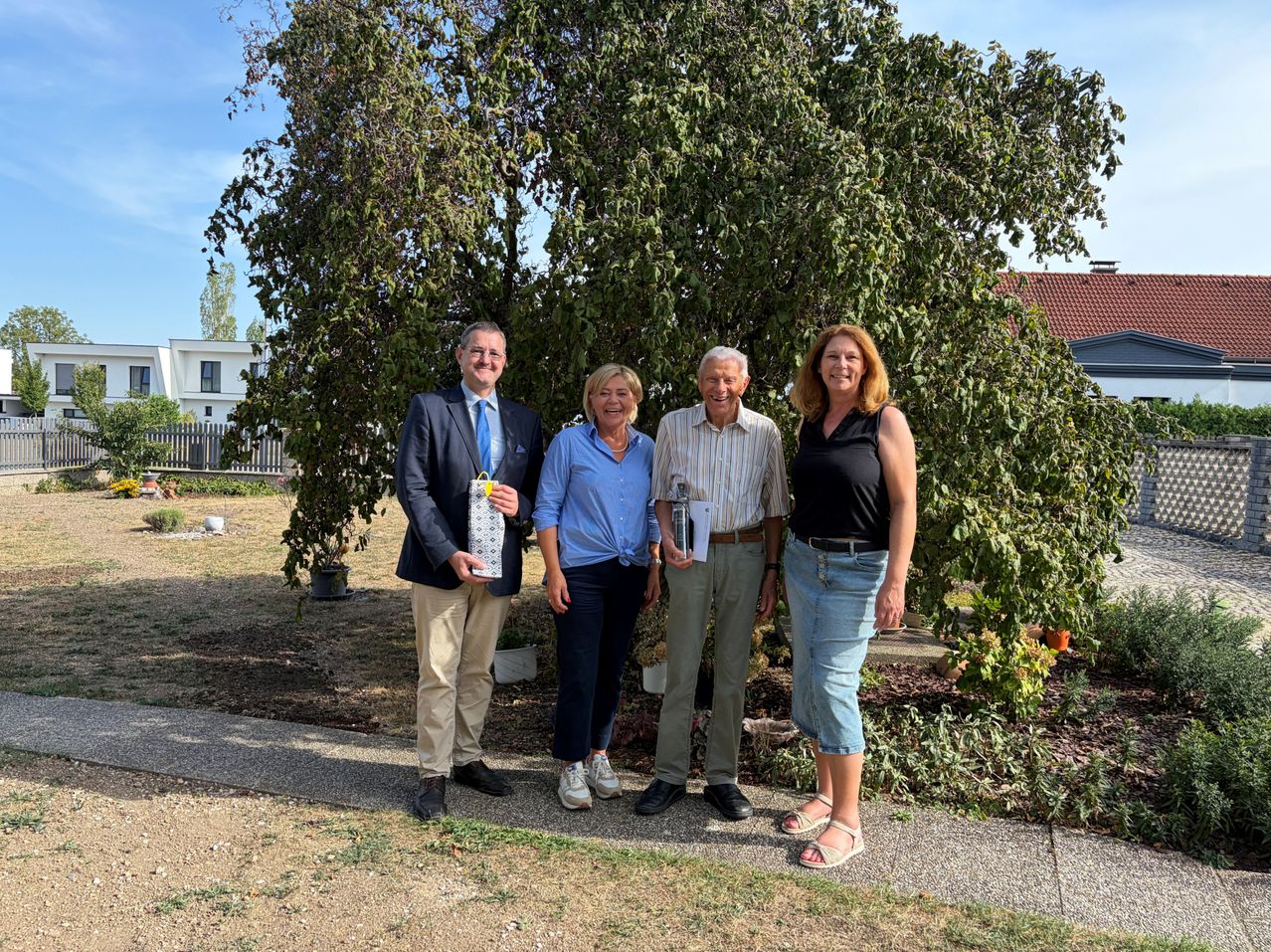 Four people are standing in front of a large tree, smiling for a photo. The man on the left holds a trophy, while the others hold papers. They are dressed casually, and a house is visible in the background.