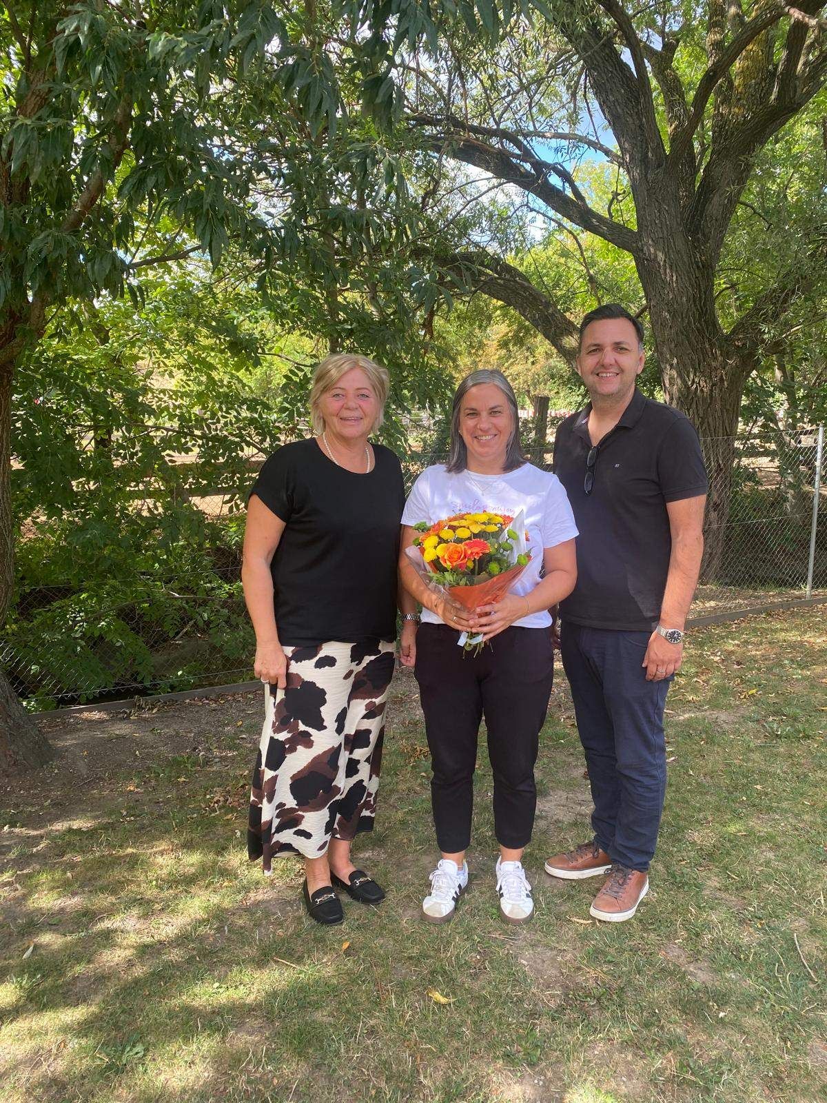 Three people stand together in a park, smiling for a photo. The woman in the middle holds a bouquet of flowers. Behind them are trees and a fence.