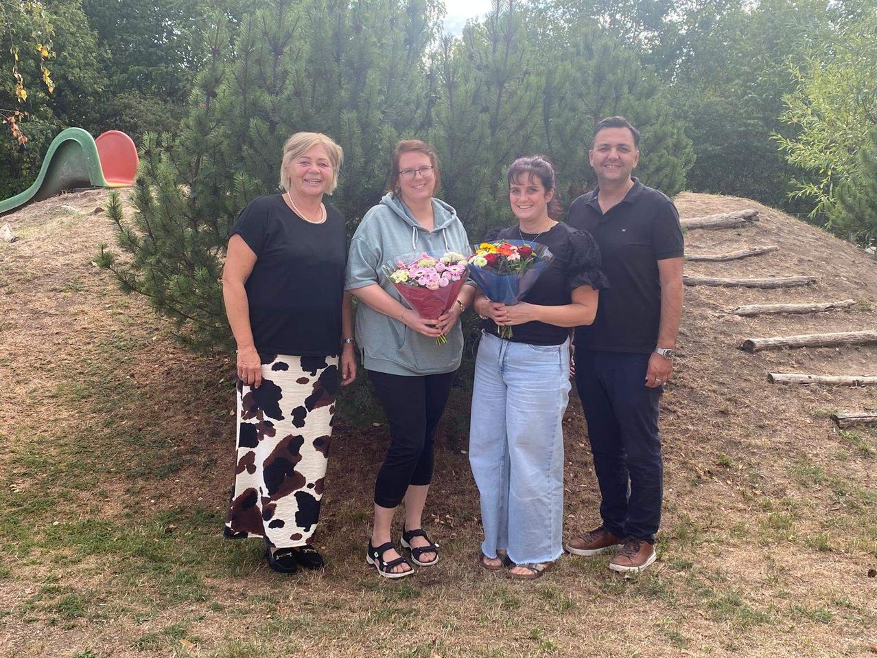 Four people stand in front of a large bush, smiling for a photo. Two women hold bouquets of flowers. The woman on the left wears a cow print skirt. The man on the right wears a watch.