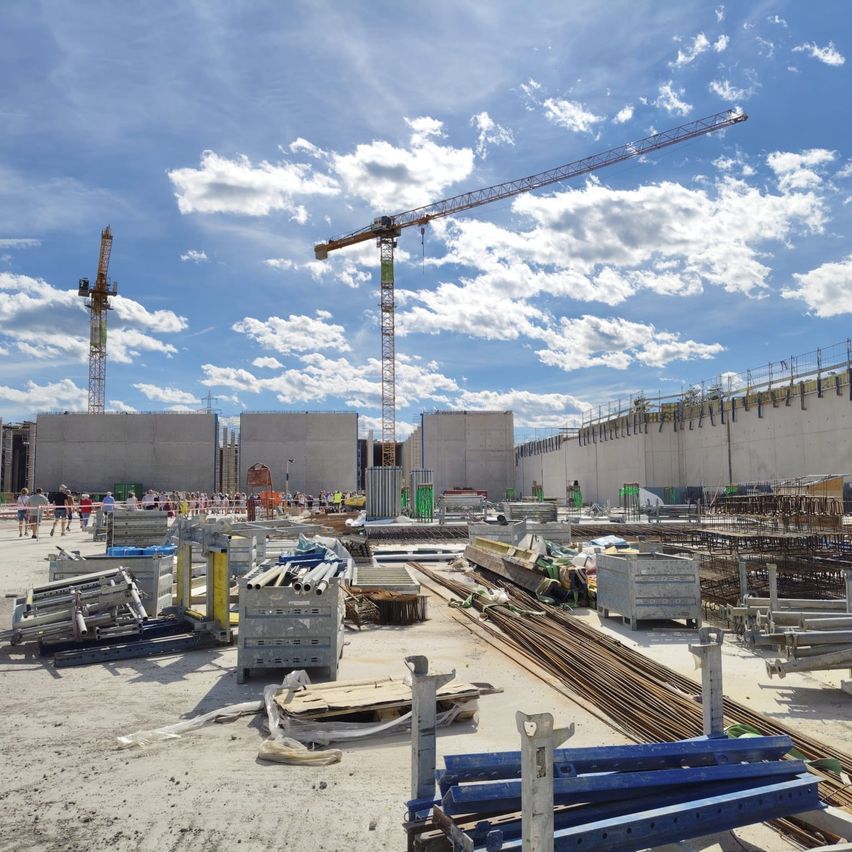 A construction site with cranes, workers, and materials under a blue sky with clouds.