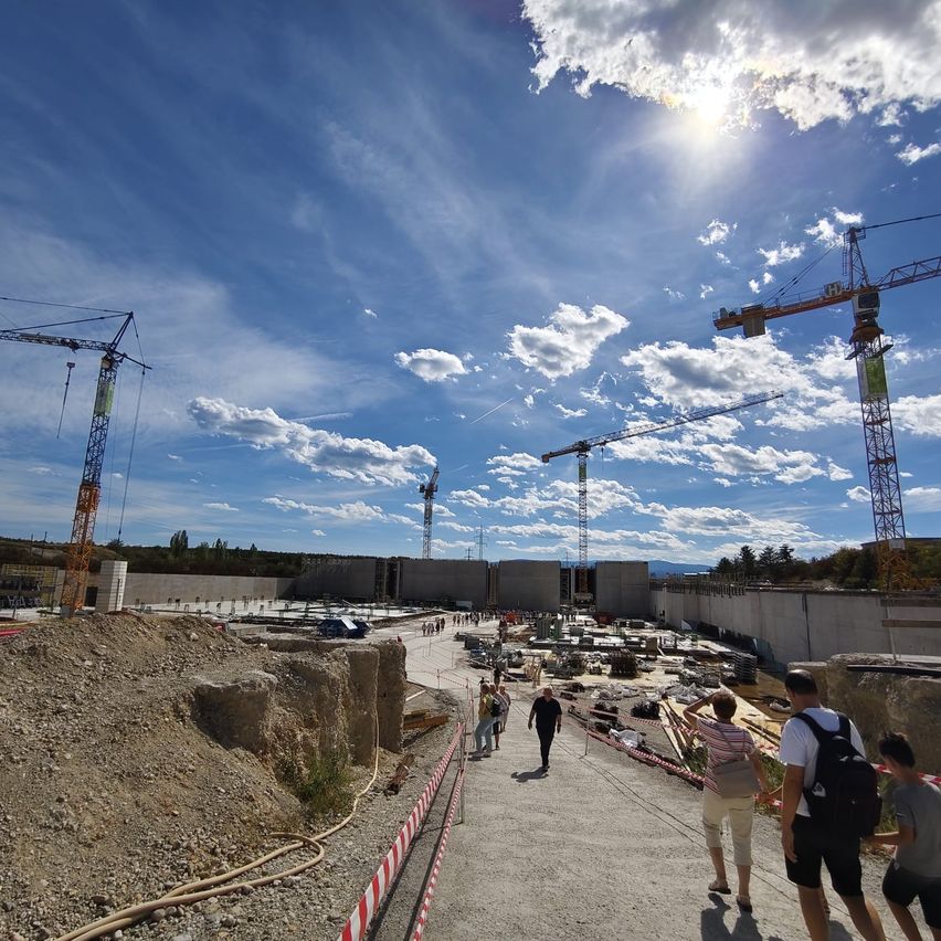 A construction site under a clear blue sky with several cranes. Workers are walking around, and some are standing near a wall.