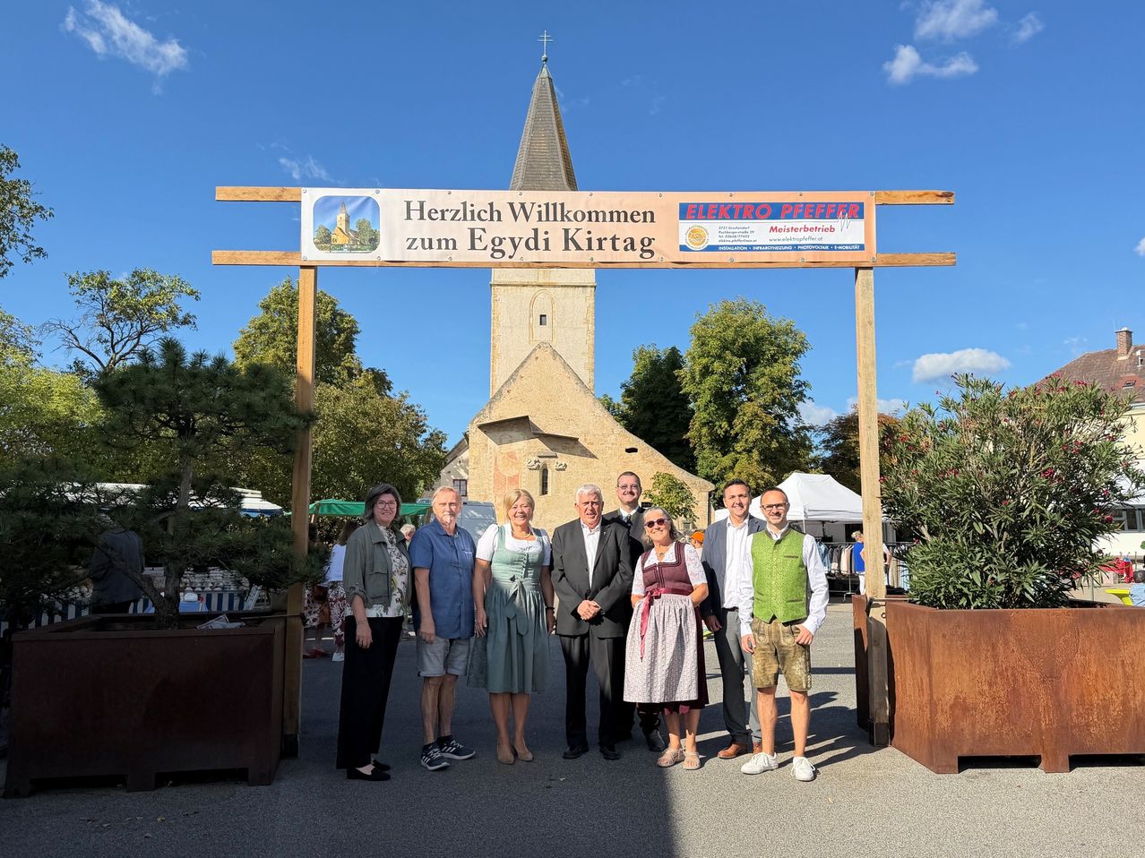 A group of people dressed in traditional attire pose for a photo in front of a church with a banner that reads Herzlich Willkommen zum Egydii Kirtag.