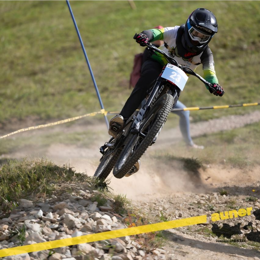 A mountain biker wearing a helmet and gloves rides over a rocky hill during a race. The number 23 is visible on the front of the bike.