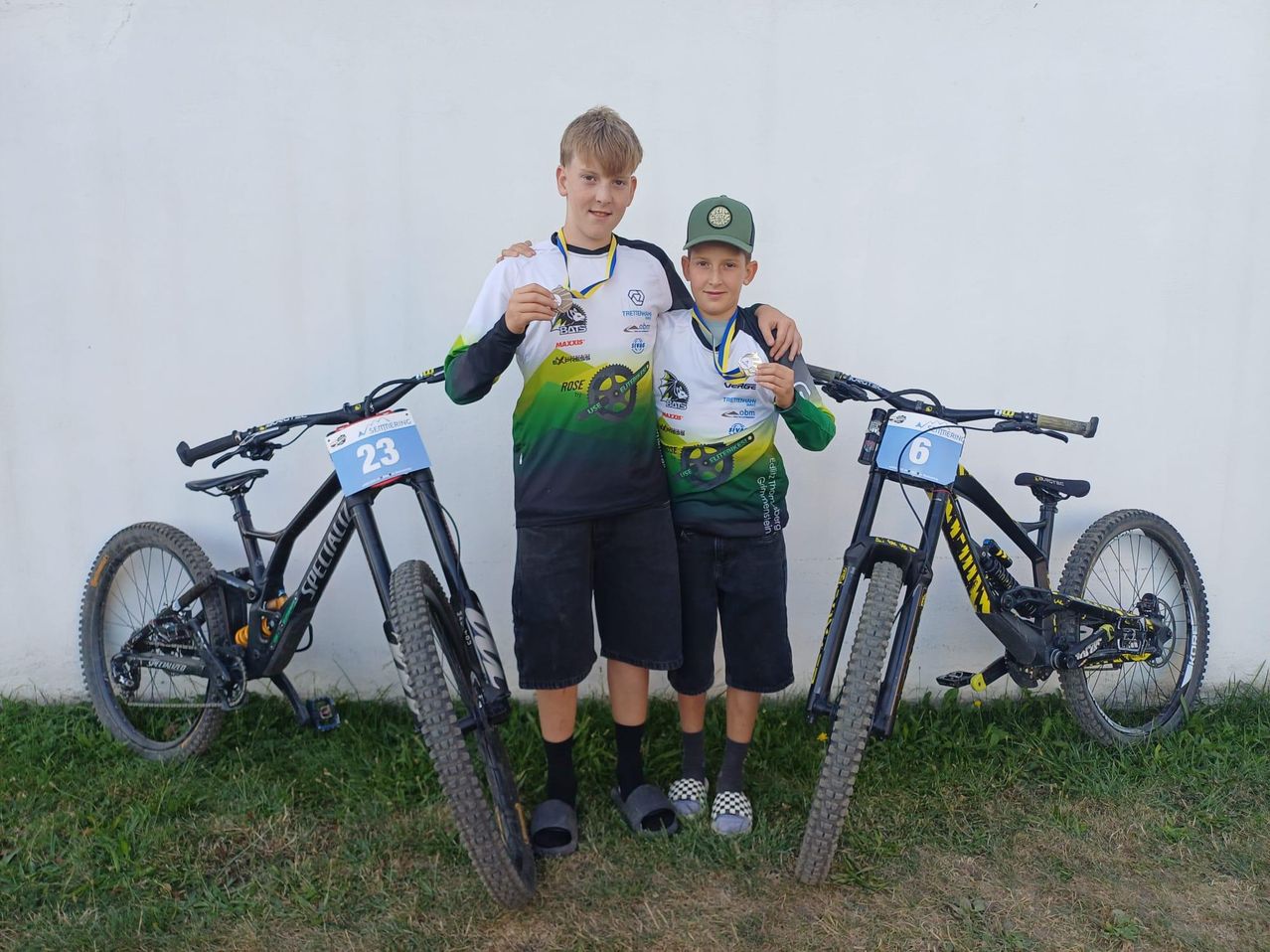 Two young boys stand next to their bikes, proudly displaying their medals. The boy on the left wears a jersey with the number 23, while the boy on the right wears a jersey with the number 6. They both have a cheerful expression.