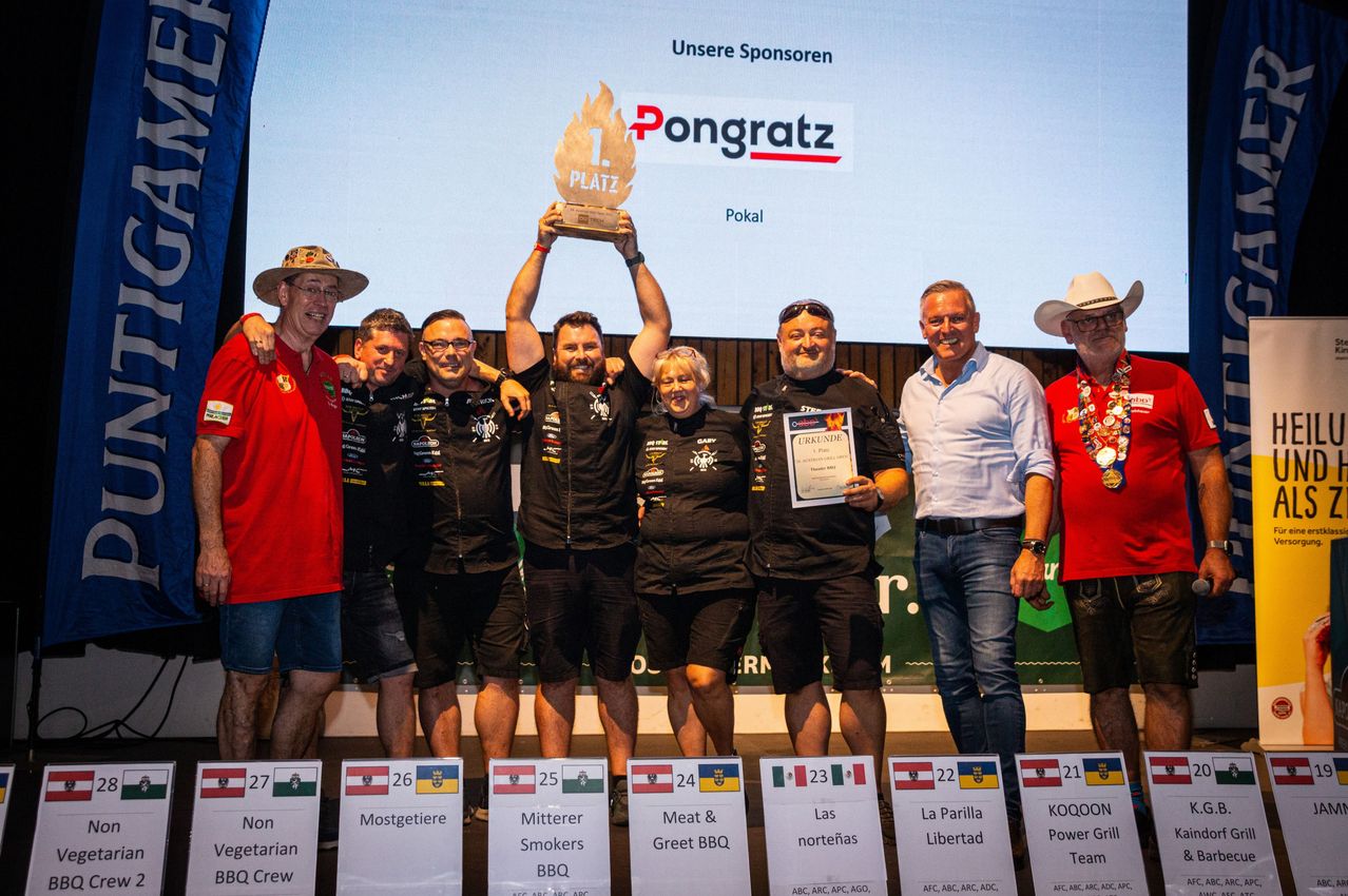A group of people stand on a stage holding a trophy and certificates. They are dressed in black and appear to be celebrating a victory. The stage has a backdrop with a sponsor's logo and a large screen behind them.