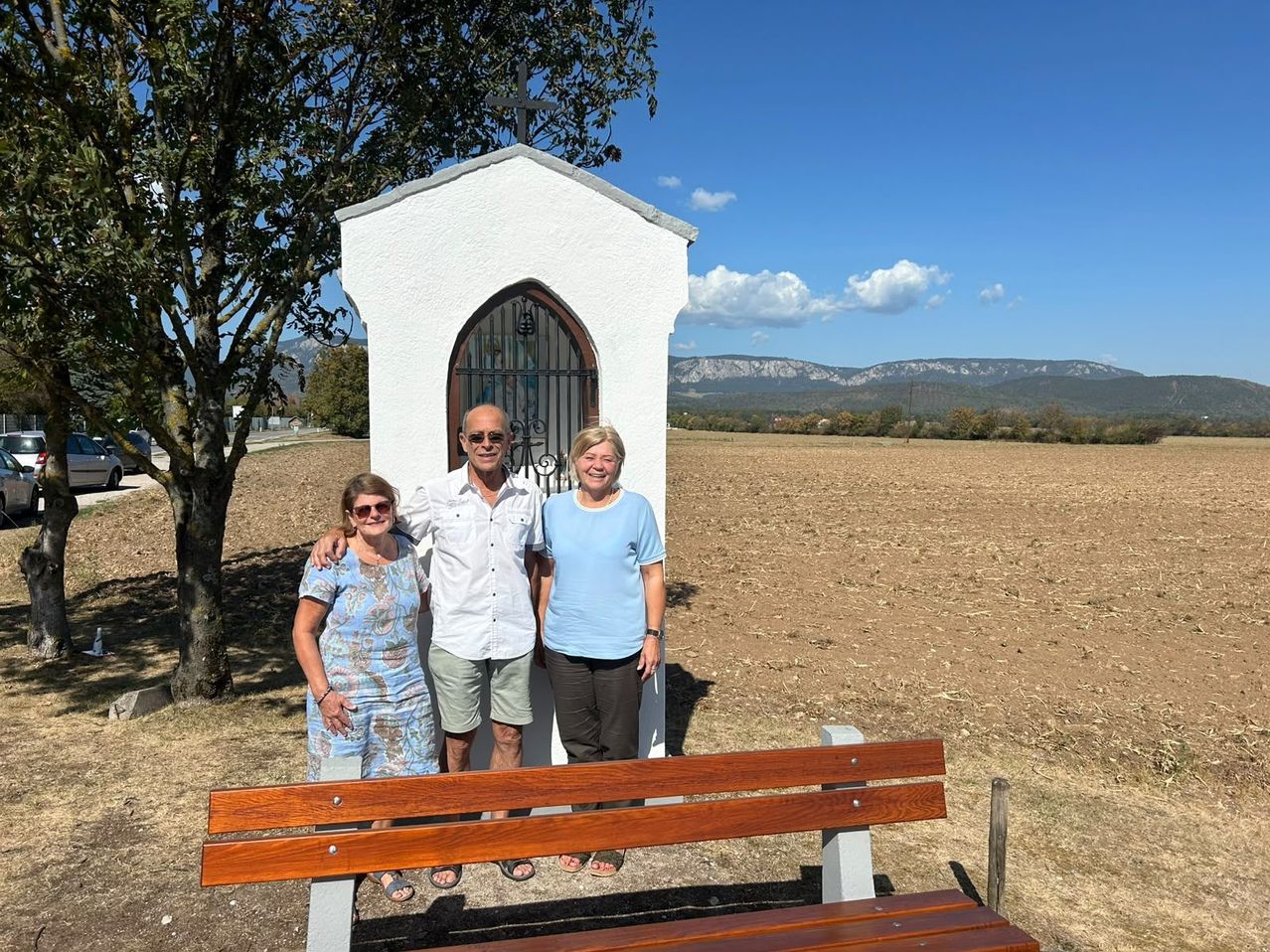 Three people stand in front of a small chapel in a rural setting, with a tree and a bench nearby. The sky is clear with a few clouds.