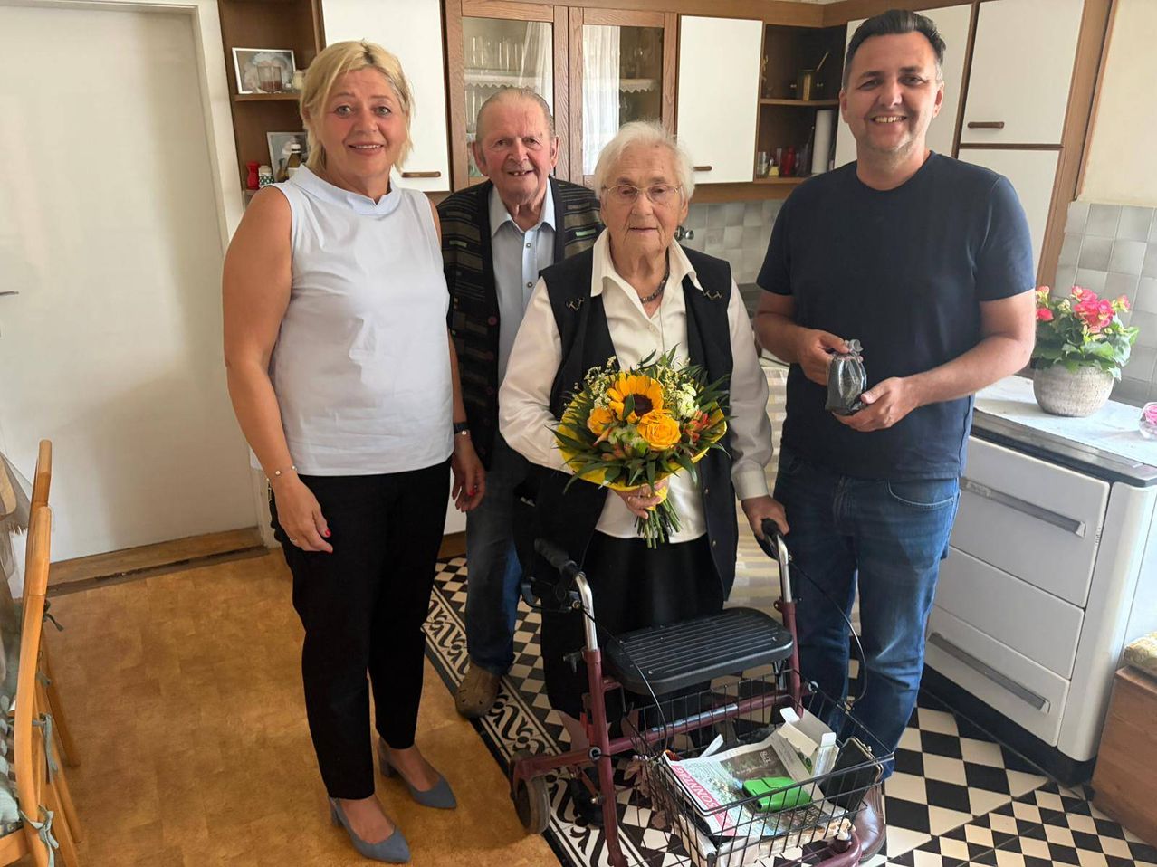 Vier Menschen stehen in einer Küche; eine ältere Frau mit einem Rollator hält einen Sonnenblumenstrauß, während ein Mann eine kleine Tasche hält. Dahinter stehen zwei andere Personen, eine in einem Weste und die andere in einem weißen Oberteil, und lächeln.