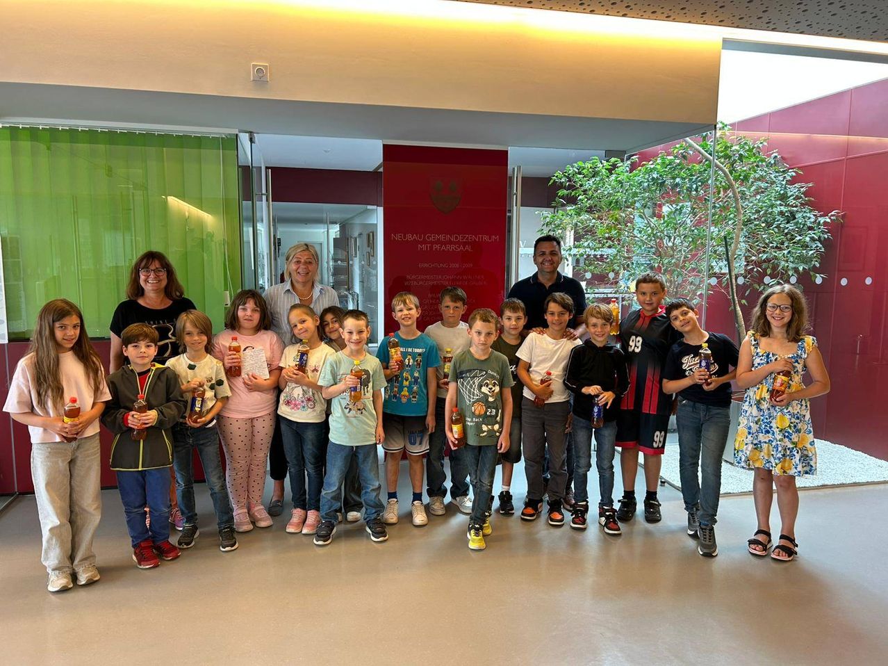 Eine Gruppe von Kindern und Erwachsenen posiert für ein Foto, hält Flaschen in der Hand und trägt verschiedene Kleidungsstücke, vor einem Gebäude mit einem Schild, das 'Neubau Gemeinschaftszentrum mit Pfarrsaal' lautet.