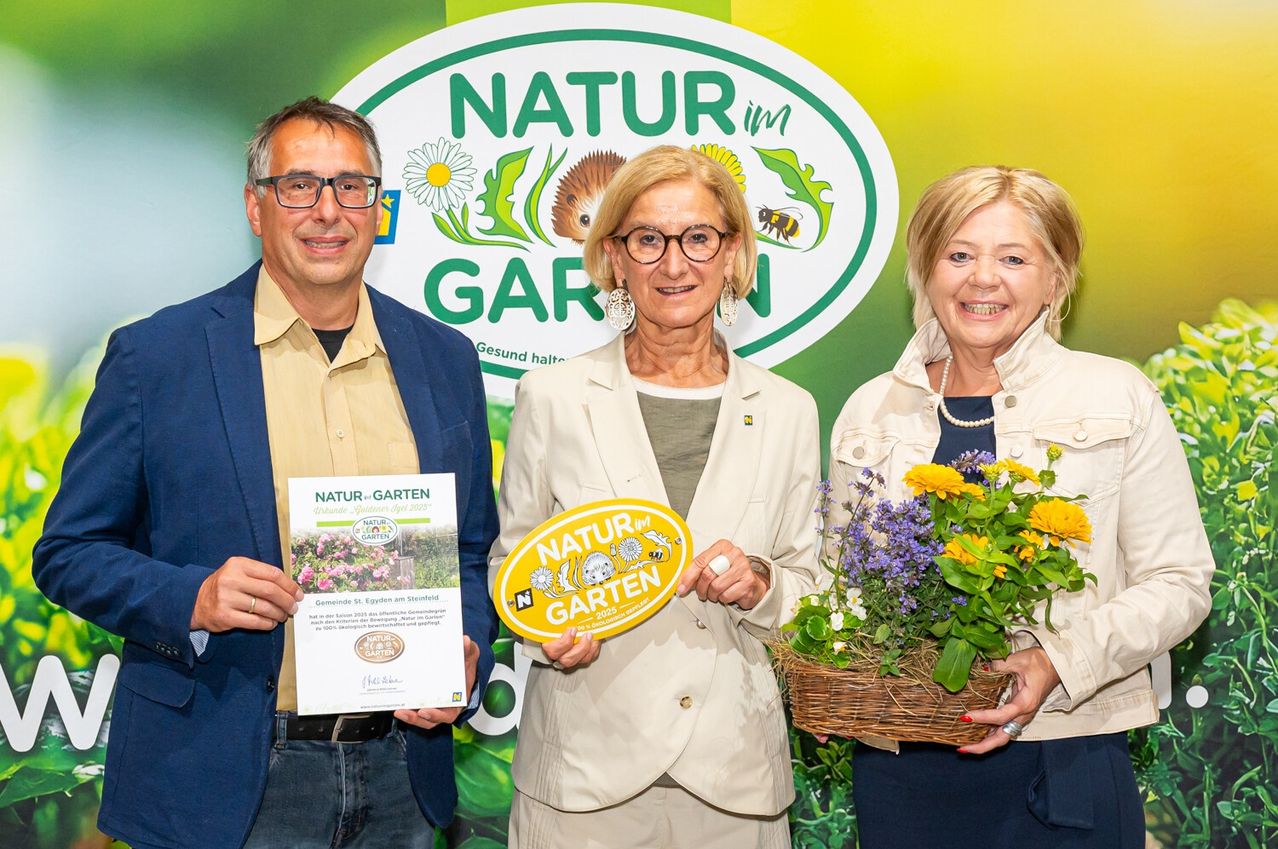 Drei Personen stehen vor einem grün-gelben Banner und lächeln. Der Mann hält ein Zertifikat, die Frau in der Mitte ein gelbes Ovalschild und die Frau auf der rechten Seite einen Blumenstrauß.
