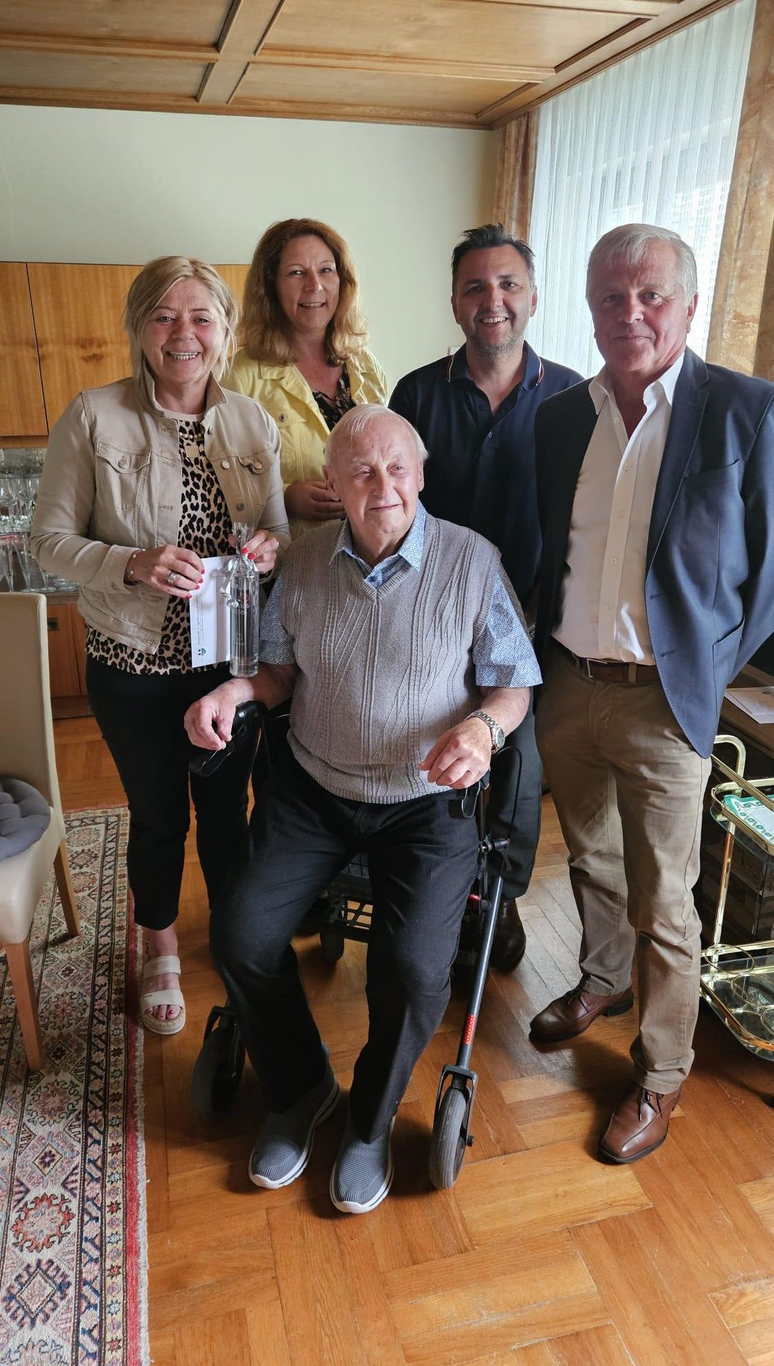 A group of people, including an elderly man in a wheelchair, are posing for a photo in a room. A woman holds a bottle and paper, while two men stand beside her. Behind them, a kitchen with a wooden cabinet is visible.