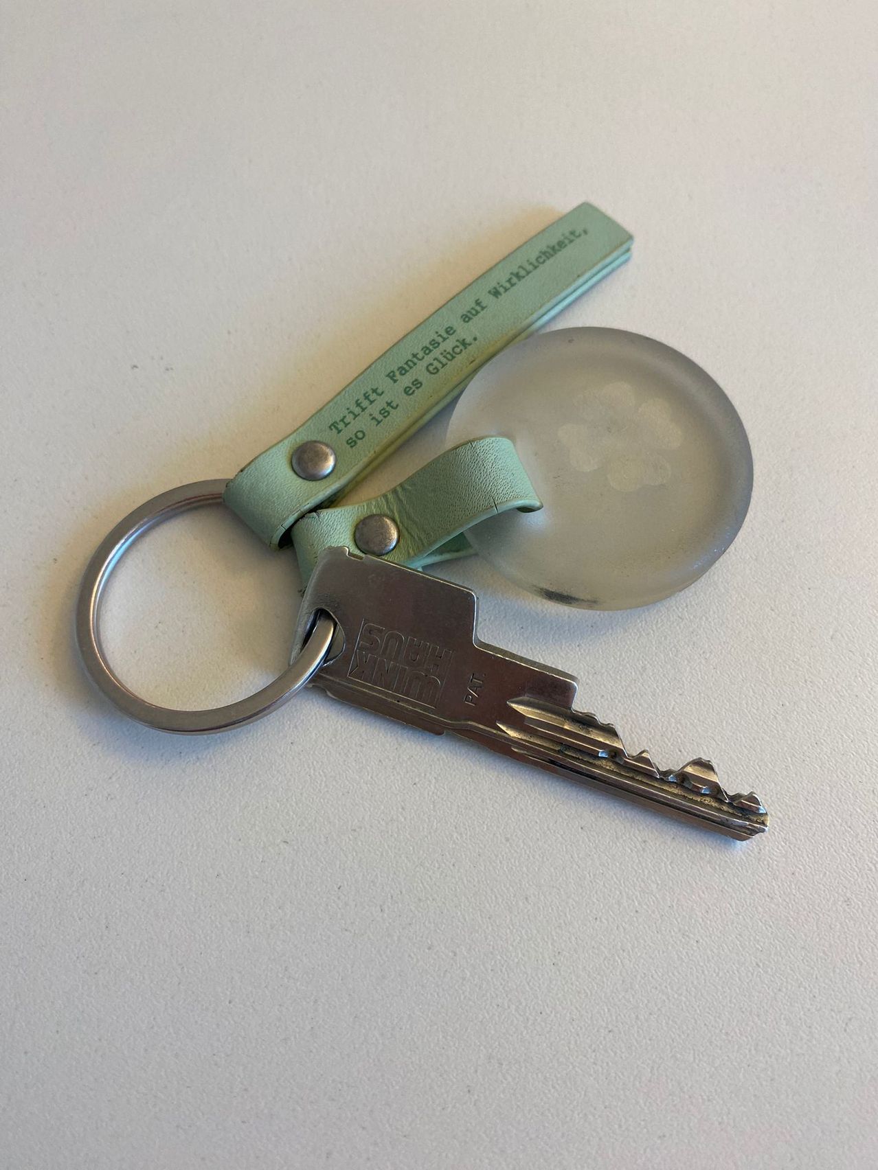 A keychain with a green strap has a silver ring and key. A glass charm with a paw print is attached. The strap reads, 'Triff Fantasie auf Wirklichkeit, so ist es Glück.'