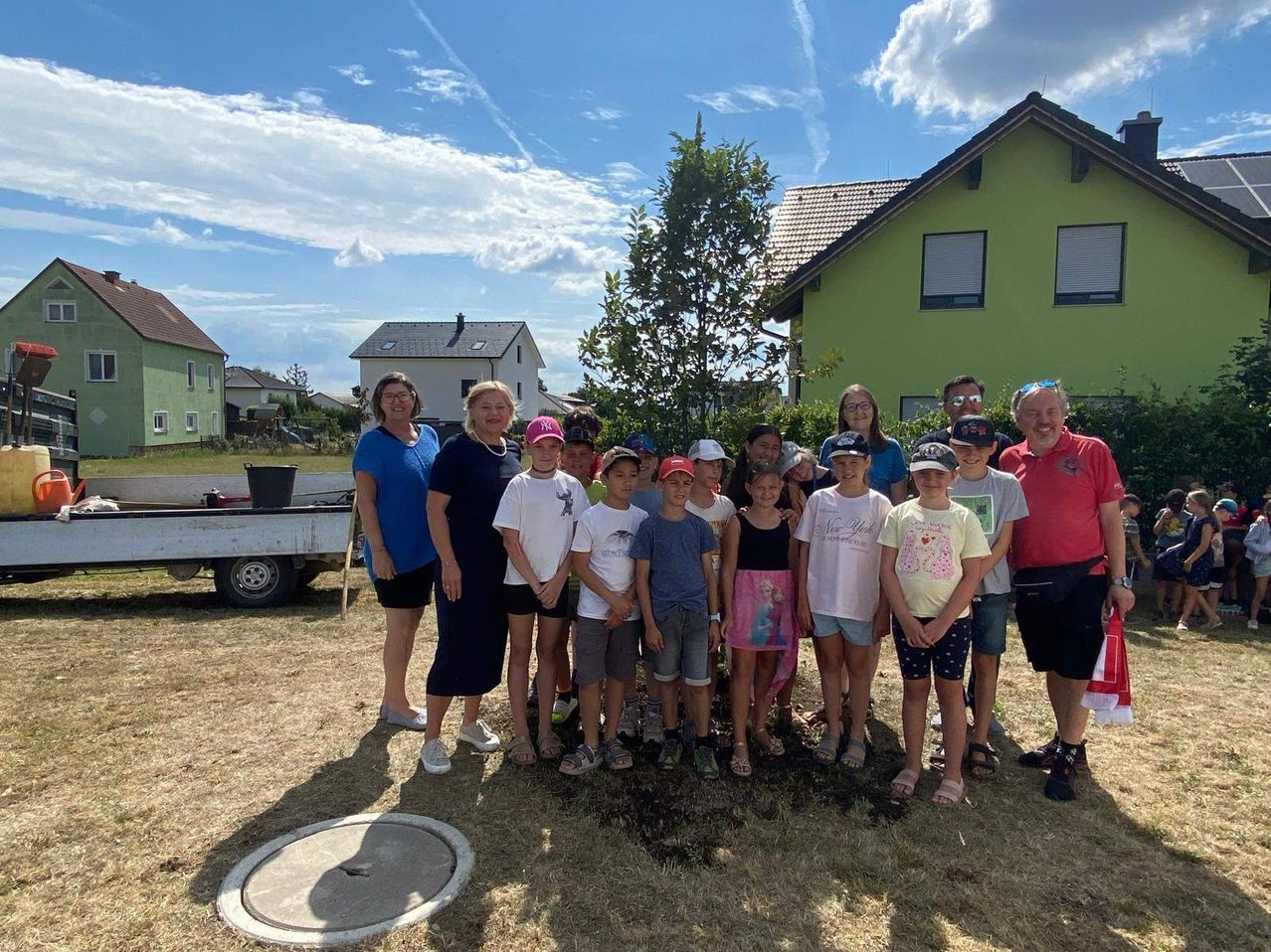 A group of people stands around a newly planted tree. They are posing for a photo with a house in the background.