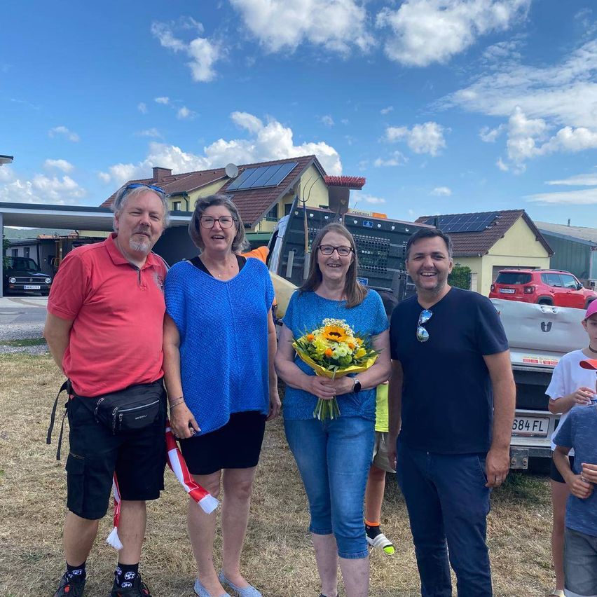 Four people stand in a field, smiling and posing for a photo. A woman holds a bouquet of flowers. Behind them are houses, a pickup truck, and a red SUV.