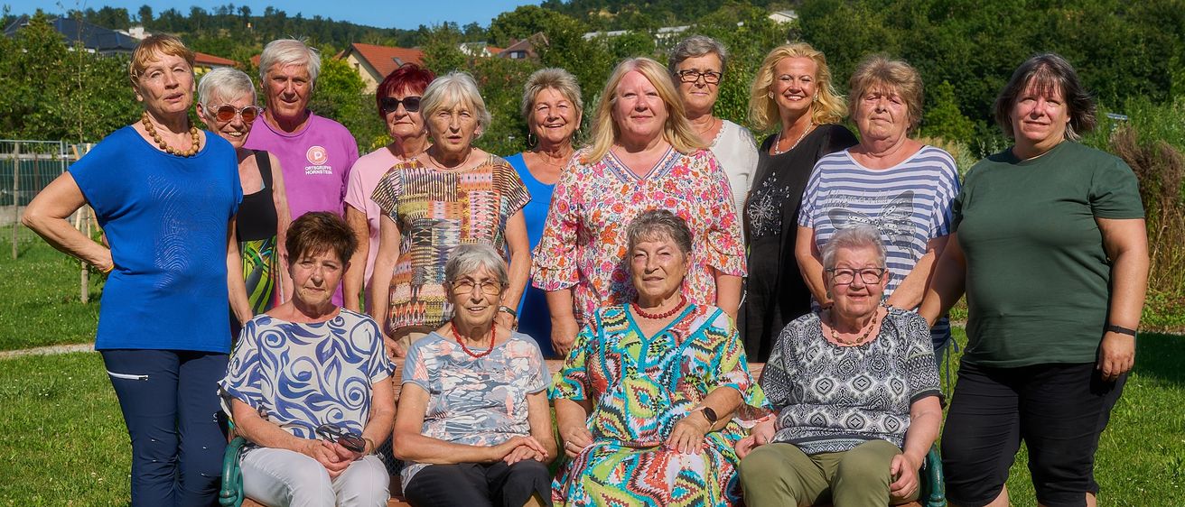 Eine Gruppe älterer Personen, einschließlich Männer und Frauen, sitzt auf einer Bank in einem Park. Sie alle lächeln und posieren für ein Foto. Der Hintergrund ist eine üppige grüne Landschaft mit Bäumen und Häusern.