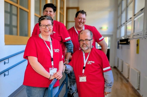Fünf Menschen in roten T-Shirts und Ausweisen lächeln und stehen in einem Flur.