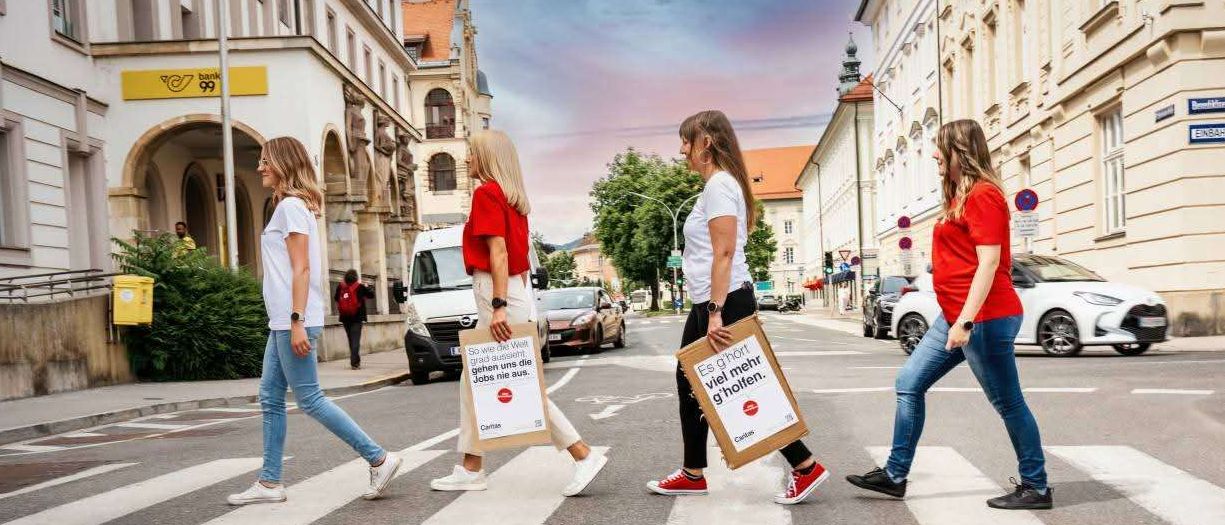 Vier Frauen, alle in roten Shirts und mit Einkaufstaschen, überqueren eine Straße. Zwei Schilder auf den Taschen lesen 'So viele Weib' und 'Es gibt mehr g'. Im Hintergrund sind Gebäude und Autos zu sehen.