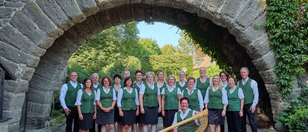 Eine Gruppe von Menschen in grünen Westen und schwarzen Röcken posiert unter einem Steinbogen mit einer Harfe auf dem Boden.