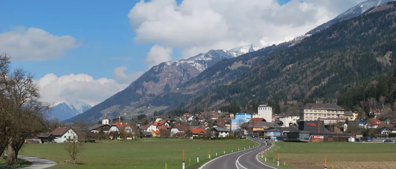 Eine Straße mit weißen Markierungen führt durch ein saftiges Feld und führt zu einem Dorf am Fuße verschneiter Berge.