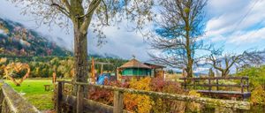 Ein Holzzaun umgibt einen farbenfrohen Garten mit Bäumen und einem kleinen grünen Gebäude, umgeben von einem Feld und Bergen im Hintergrund.