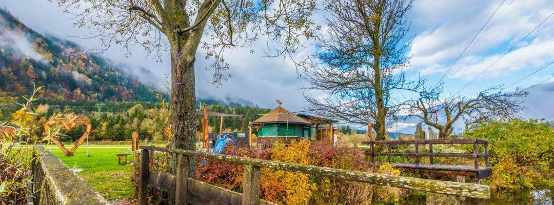 Ein Holzzaun umgibt einen farbenfrohen Garten mit Bäumen und einem kleinen grünen Gebäude, umgeben von einem Feld und Bergen im Hintergrund.