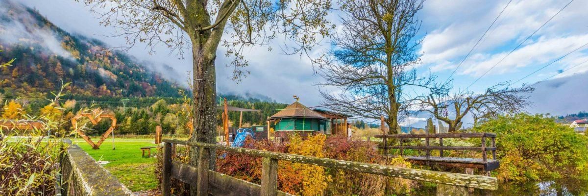 Ein Holzzaun umgibt einen farbenfrohen Garten mit Bäumen und einem kleinen grünen Gebäude, umgeben von einem Feld und Bergen im Hintergrund.