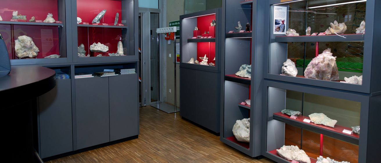 Ein Raum mit Holzboden, der verschiedene Mineralproben auf Regalen ausstellt. Die Regale sind grau mit roten Akzenten. Es gibt Bücher und zusätzliche Objekte in den unteren Schubladen.