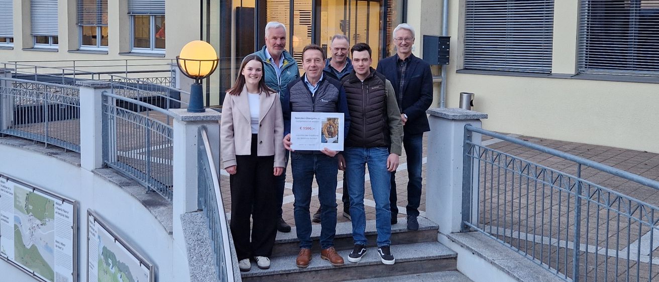 Fünf Personen stehen auf einer Treppe vor einem Gebäude mit einem Schild, das 'Gemeinde Mühldorf' steht. Sie halten ein Zertifikat.