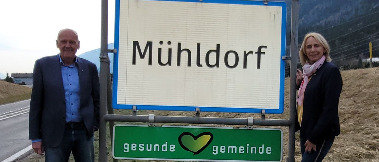 Zwei Personen, ein Mann und eine Frau, stehen neben einem Straßenschild mit blauem Rand. Das Schild trägt den Namen 'Muhldorf' in Schwarz. Darunter befindet sich ein grünes Schild mit einem Herz und den Worten 'gesunde gemeinde'. Hinter ihnen liegt ein Grasfeld und ein Berg.