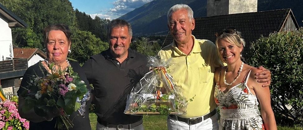 Four people stand in a garden with flowers and a church tower in the background. The man on the right holds a bouquet of flowers, while the man on the left holds a bouquet of flowers and a ribbon.