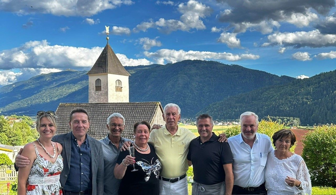 Eine Gruppe von sechs Personen, die lächelnd und für ein Foto vor einer Kirche mit einem Turm und einem Berg im Hintergrund posiert.