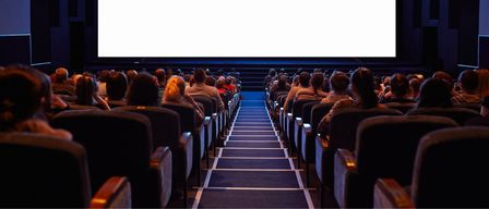 An audience sits in rows of seats facing a large blank screen in a theater, illuminated by blue lighting.