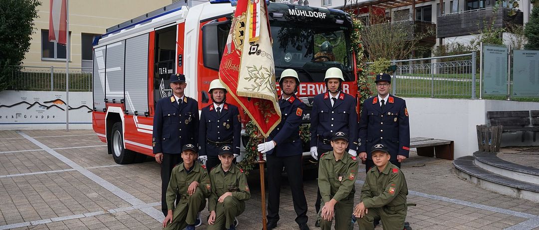 Eine Gruppe von Feuerwehrleuten in Uniform steht vor einem Feuerwehrauto mit dem Wort Muhldorf. Sie posieren für ein Foto, einige sitzend und andere stehend, halten eine Fahne. Dahinter befindet sich ein Gebäude mit Balkon.