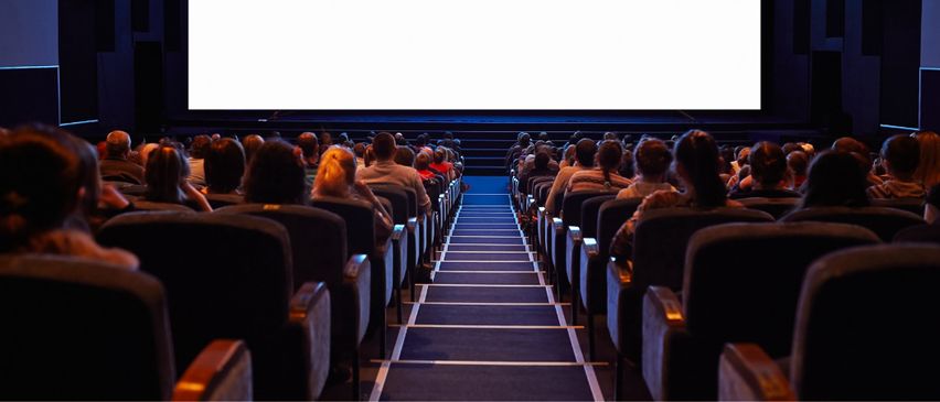 Ein überfülltes Kino mit einer großen Leinwand, Reihen von Stühlen und blauen Teppichgängen. Das Publikum sieht einen Film.