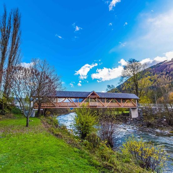 Eine Holzbrücke erstreckt sich über einen Fluss mit üppigem Grün und Bergen im Hintergrund unter einem blauen Himmel mit verstreuten Wolken.
