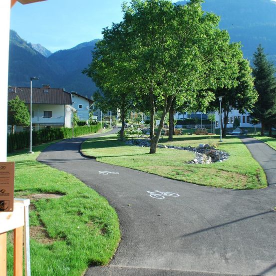 Ein gepflasterter Weg schlängelt sich durch einen Park mit üppigem Gras und Bäumen und führt zu einem Gebäude unter einer Bergkulisse. Fahrradsymbole markieren den Weg.